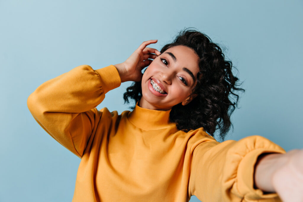 Beautiful young woman with traditional metal braces smiling and tucking her hair behind her ear
